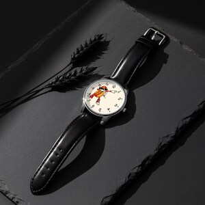 May include: A wristwatch with a black leather strap and a silver-tone metal case. The watch face is off-white with black numerals and features a central graphic of a cartoon hot dog character wearing sunglasses and sneakers in a dabbing pose.
