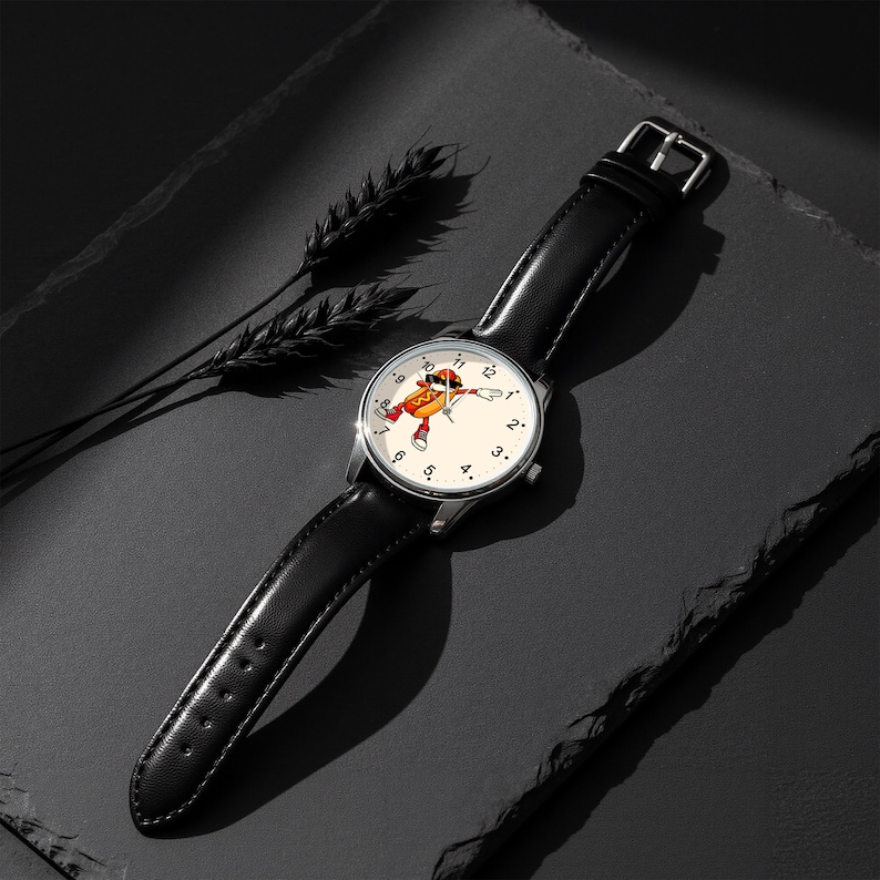 May include: A wristwatch with a black leather strap and a silver-tone metal case. The watch face is off-white with black numerals and features a central graphic of a cartoon hot dog character wearing sunglasses and sneakers in a dabbing pose.