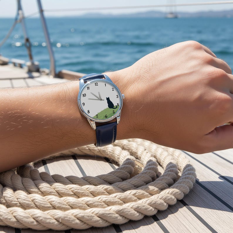 Op de afbeelding: Een polshorloge met een witte wijzerplaat en zwarte cijfers en wijzers. De wijzerplaat heeft een groene heuvel met een zwart katten silhouet. Het horloge heeft een zilveren kast en een marineblauwe leren band. Het horloge zit om de pols van een persoon.