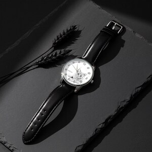 May include: A silver wristwatch with a black leather band. The watch face features a detailed illustration of a cat and the numbers 12, 3, and 9. The watch is set against a dark background with a sprig of wheat.