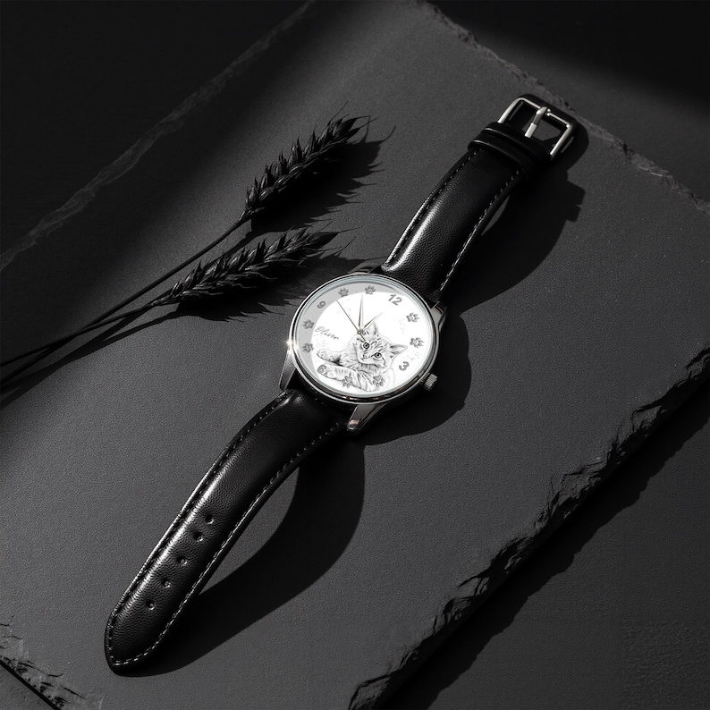 May include: A silver wristwatch with a black leather band. The watch face features a detailed illustration of a cat and the numbers 12, 3, and 9. The watch is set against a dark background with a sprig of wheat.