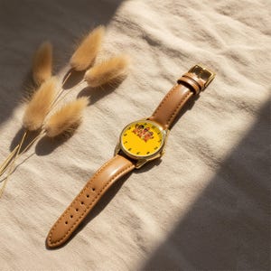 May include: A wristwatch with a gold-tone metal case and a tan leather strap. The watch face is bright yellow and features a graphic illustration of three hot dog characters wearing sunglasses. The numbers one through twelve are printed in black.