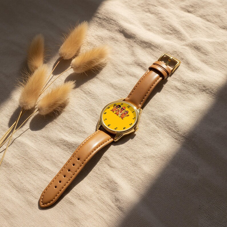 May include: A wristwatch with a gold-tone metal case and a tan leather strap. The watch face is bright yellow and features a graphic illustration of three hot dog characters wearing sunglasses. The numbers one through twelve are printed in black.