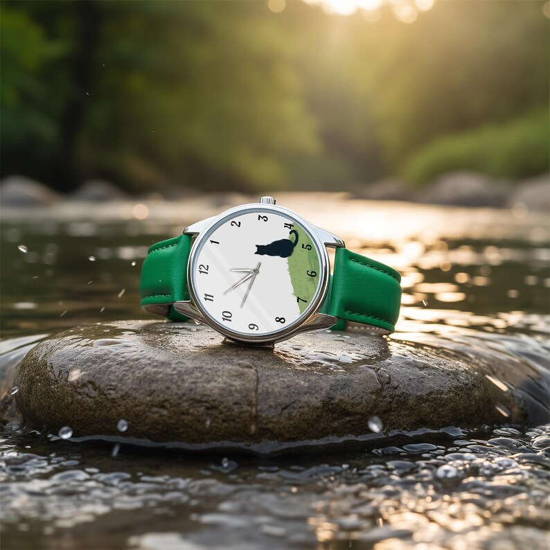 Op de afbeelding: Een zilveren polshorloge met een witte wijzerplaat met zwarte cijfers en een zwart katten silhouet op een groen veld. Het horloge heeft een groene leren band. Het horloge ligt op een rots in een beek.