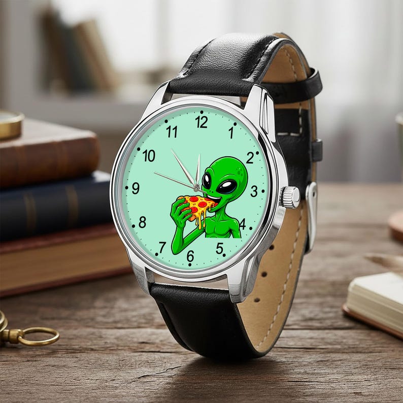 May include: A wristwatch with a black leather strap and a silver-tone metal case. The watch face is light green and features a graphic illustration of a green alien eating a slice of pepperoni pizza, with black numerals marking the hours.