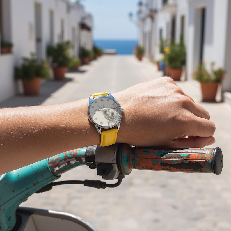 Pu&ograve; includere: Un orologio da polso con cinturino giallo e cassa color argento. Il quadrante dell'orologio presenta un'immagine in bianco e nero di un cane. L'orologio &egrave; indossato su un polso, con un manubrio di scooter in primo piano.