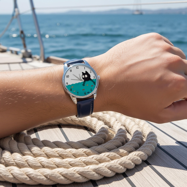 Pu&ograve; includere: Orologio da polso con quadrante bianco con illustrazione di un gatto nero. L'orologio ha un cinturino in pelle blu e una cassa color argento. Il gatto &egrave; raffigurato in acqua con pesci. L'orologio &egrave; al polso.