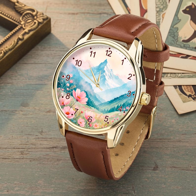 May include: A wristwatch with a brown leather strap and a gold-tone metal case. The watch face features a watercolor-style illustration of a mountain landscape with pink flowers in the foreground, gold-tone hands, and black numerical hour markers.
