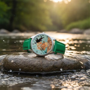 Puede incluir: Un reloj de pulsera plateado con una correa de cuero verde. La esfera del reloj presenta un gato negro, flores y un jarr&oacute;n sobre un fondo azul claro. El reloj est&aacute; sobre una roca en un arroyo.