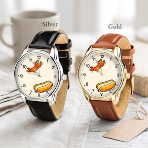 May include: Two wristwatches with cream-colored faces featuring a cartoon hot dog character and a hot dog illustration. One watch has a silver-tone metal case with a black leather strap, and the other has a gold-tone metal case with a brown leather strap.