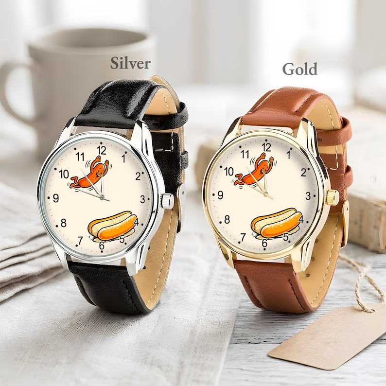 May include: Two wristwatches with cream-colored faces featuring a cartoon hot dog character and a hot dog illustration. One watch has a silver-tone metal case with a black leather strap, and the other has a gold-tone metal case with a brown leather strap.