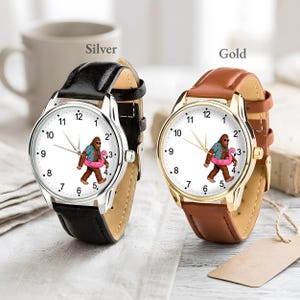 May include: Two analog wristwatches with white faces featuring a graphic of a brown creature wearing a floral shirt and holding a pink flamingo float. One watch has a silver-tone case with a black leather strap, and the other has a gold-tone case with a brown leather strap.