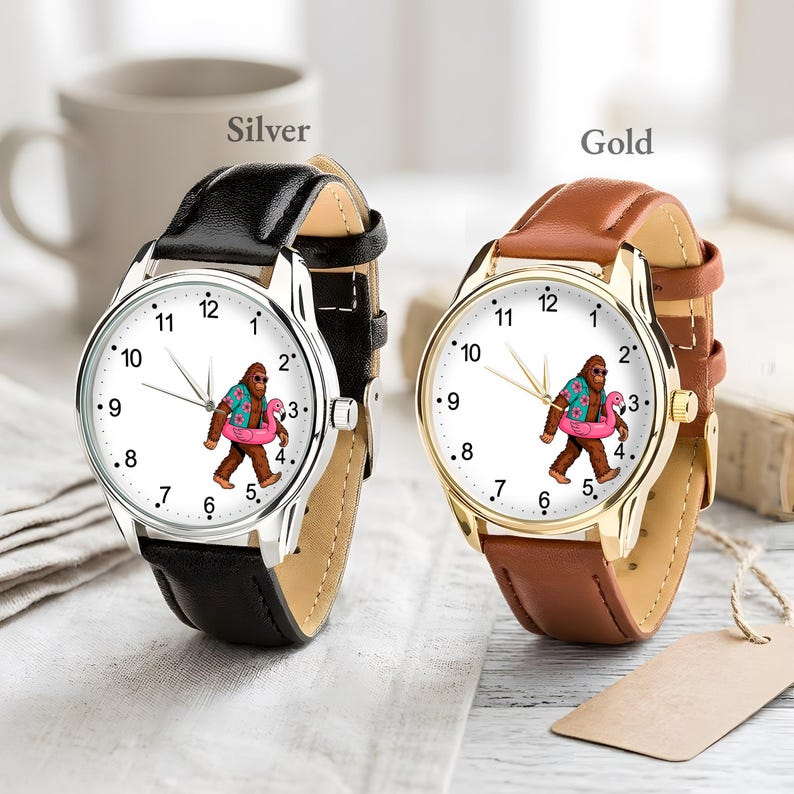 May include: Two analog wristwatches with white faces featuring a graphic of a brown creature wearing a floral shirt and holding a pink flamingo float. One watch has a silver-tone case with a black leather strap, and the other has a gold-tone case with a brown leather strap.