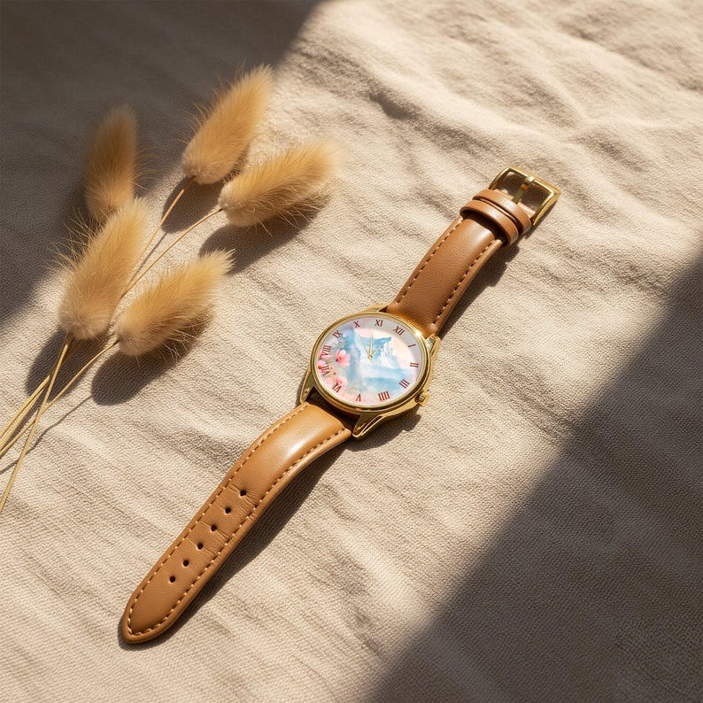 K&ouml;nnte beinhalten: Eine goldfarbene Armbanduhr mit hellbraunem Lederarmband. Das Zifferblatt zeigt ein Aquarelldesign in Rosa und Blau. R&ouml;mische Ziffern markieren die Stunden. Die Uhr liegt vor einem neutralen Hintergrund.