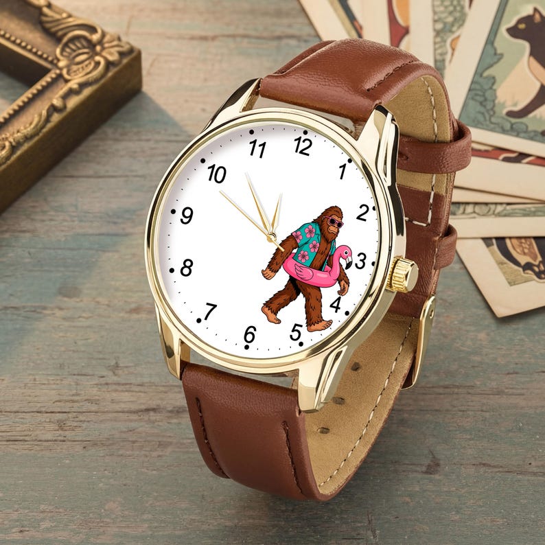 May include: A wristwatch with a gold-tone metal case and a brown leather strap. The white watch face features black numerals and a graphic of a brown Bigfoot wearing a floral shirt and sunglasses while carrying a pink flamingo pool float.