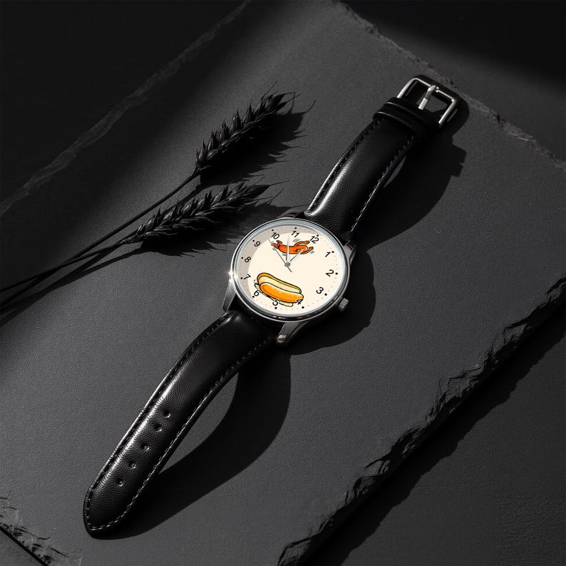 May include: A wristwatch with a black leather strap and a silver-tone case. The watch face is off-white with black numerals and features a graphic illustration of a hot dog and a small flying creature in the center, with thin black hands.