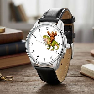 May include: A wristwatch with a black leather strap and a silver-tone metal case. The white watch face features black Arabic numerals and a central graphic illustration of a brown Bigfoot creature riding on the back of a green Tyrannosaurus Rex.