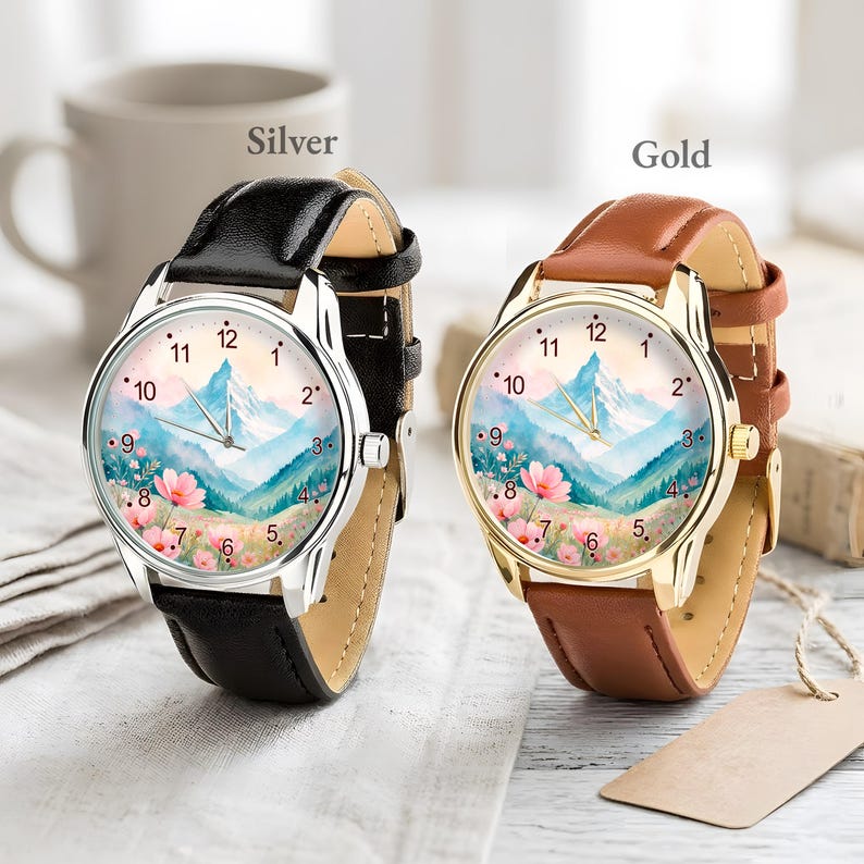 May include: Two analog wristwatches featuring a watercolor mountain and floral landscape on the dial. One watch has a silver-tone case with a black leather strap, and the other has a gold-tone case with a brown leather strap. Both have numeric markers.
