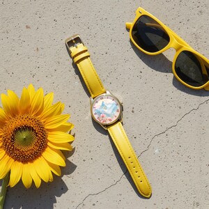 K&ouml;nnte beinhalten: Eine gelbe Uhr mit einem goldfarbenen Geh&auml;use und einem passenden gelben Lederarmband. Das Zifferblatt der Uhr zeigt ein Aquarelldesign mit Bergen und Blumen. Eine gelbe Sonnenbrille liegt daneben, zusammen mit einer leuchtend gelben Sonnenblume.