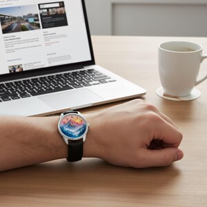 May include: A wristwatch with a black leather band and a silver-colored case. The watch face features a colorful mountain landscape with blue, orange, and pink hues. The watch is on a person's wrist, near a laptop and a coffee cup.