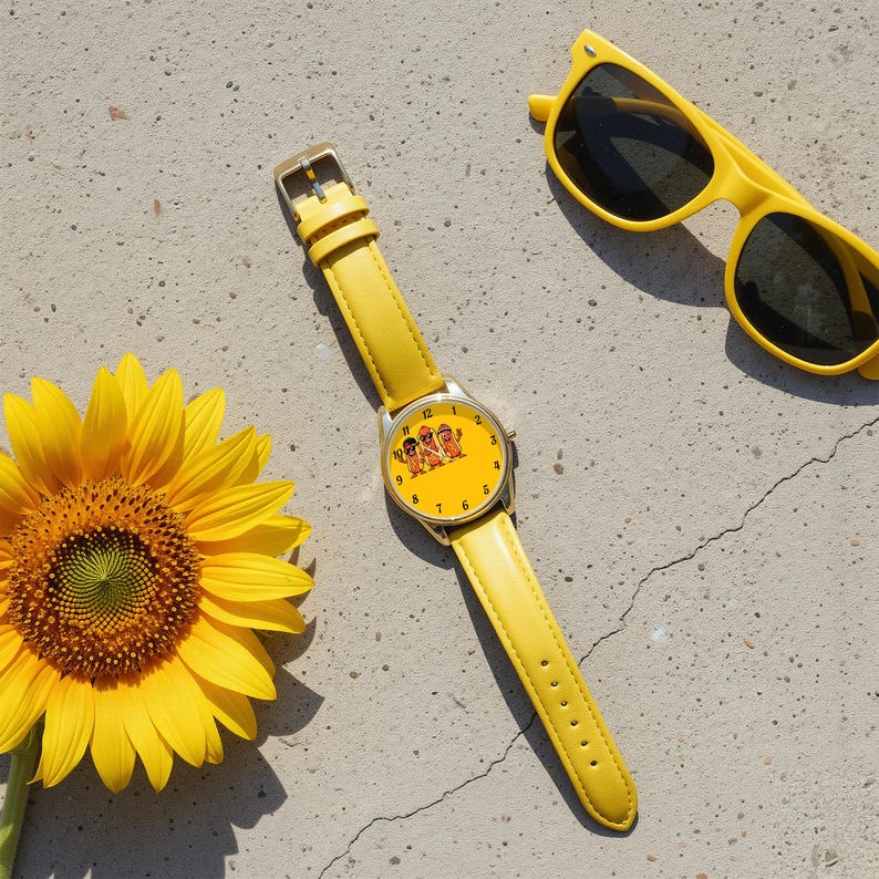 May include: A yellow wristwatch with a matching yellow leather strap and a round face featuring a graphic of three hot dogs. The watch is positioned next to a bright yellow sunflower and a pair of yellow-framed sunglasses with dark lenses.
