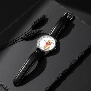 Puede incluir: Un reloj de pulsera plateado con una correa de cuero negro. La esfera del reloj presenta una ilustraci&oacute;n en acuarela de un golden retriever con un pa&ntilde;uelo rojo. El reloj est&aacute; sobre un fondo oscuro.