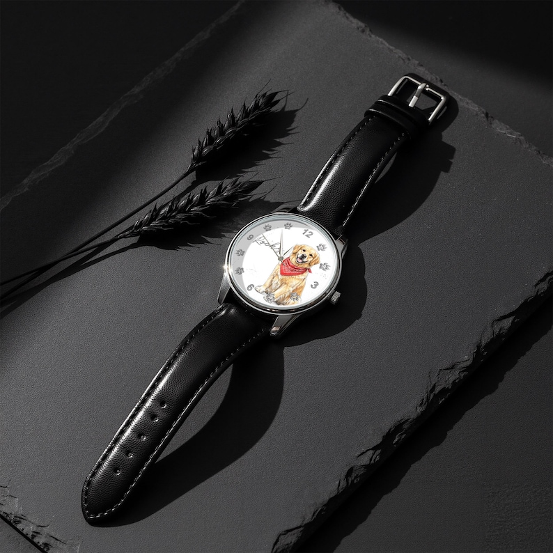 Puede incluir: Un reloj de pulsera plateado con una correa de cuero negro. La esfera del reloj presenta una ilustraci&oacute;n en acuarela de un golden retriever con un pa&ntilde;uelo rojo. El reloj est&aacute; sobre un fondo oscuro.