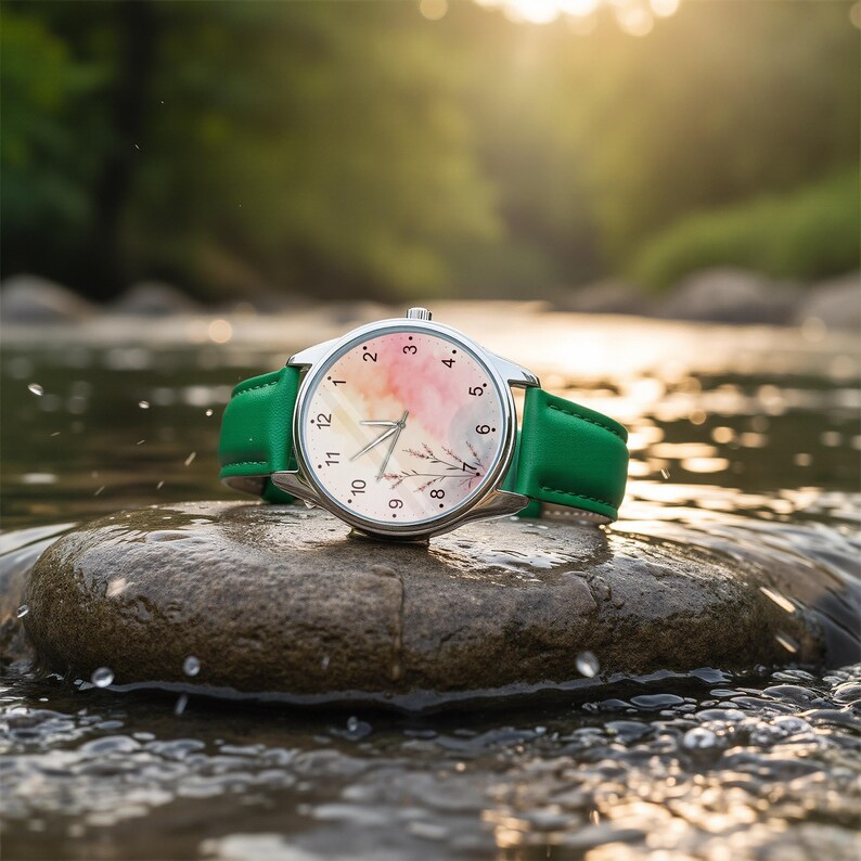 May include: A wristwatch with a silver-toned circular case and a vibrant green leather strap. The watch face features a soft watercolor-style background in shades of pink and yellow, accented with a delicate botanical illustration and black numerals.