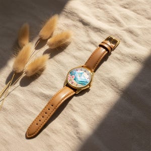 May include: A wristwatch with a gold-tone metal case and a tan leather strap. The watch face features a colorful illustration of a landscape with pink flowers, blue mountains, and a green field, marked with black numbers from one to twelve.