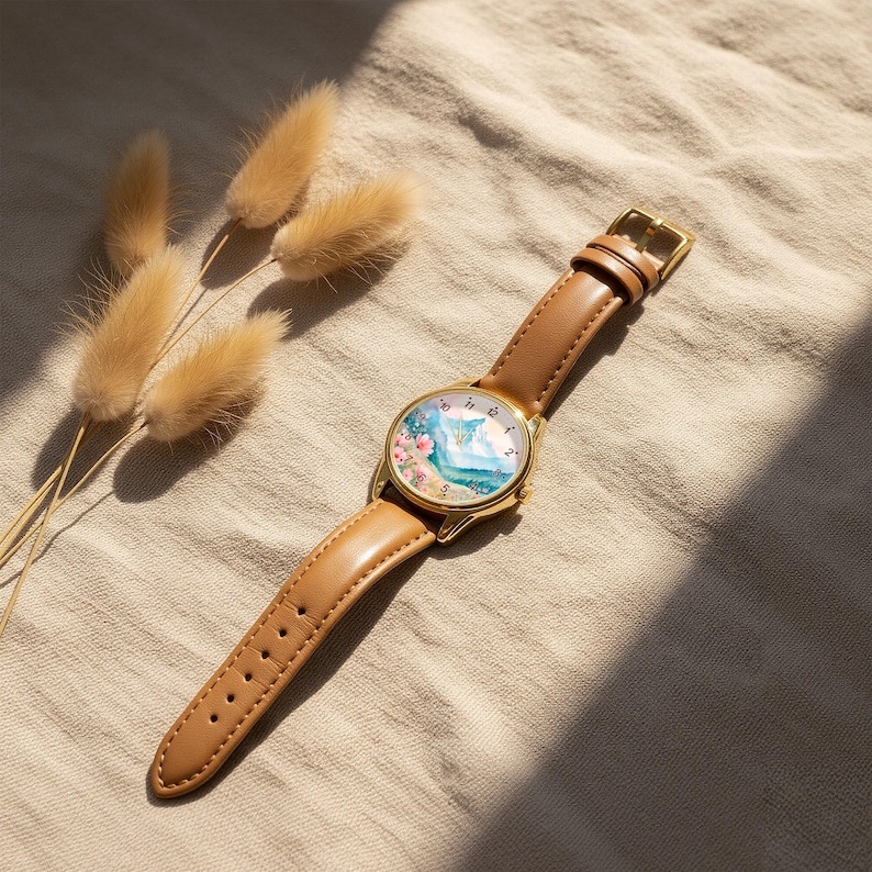 May include: A wristwatch with a gold-tone metal case and a tan leather strap. The watch face features a colorful illustration of a landscape with pink flowers, blue mountains, and a green field, marked with black numbers from one to twelve.