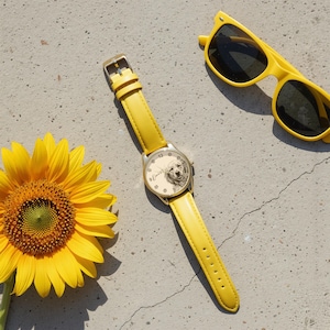 Pu&ograve; includere: Un orologio giallo con cassa color oro e cinturino in pelle giallo. Il quadrante dell'orologio presenta un ritratto di cane e il nome "Daisy". Occhiali da sole gialli e un girasole sono anche nell'immagine.