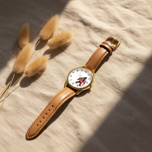 May include: A wristwatch with a gold-tone metal case and a tan leather strap. The white watch face features black numerals and a central graphic of a brown creature wearing a blue shirt and a pink inner tube, with gold-tone hour and minute hands.
