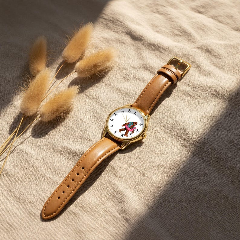 May include: A wristwatch with a gold-tone metal case and a tan leather strap. The white watch face features black numerals and a central graphic of a brown creature wearing a blue shirt and a pink inner tube, with gold-tone hour and minute hands.