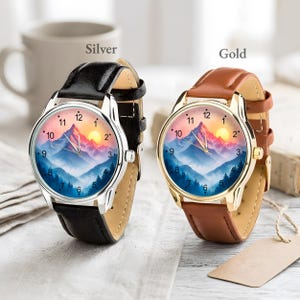 May include: Two wristwatches with mountain landscape dials. One watch has a silver case and black leather strap, the other has a gold case and brown leather strap. The watch faces feature a mountain range with a sunset.