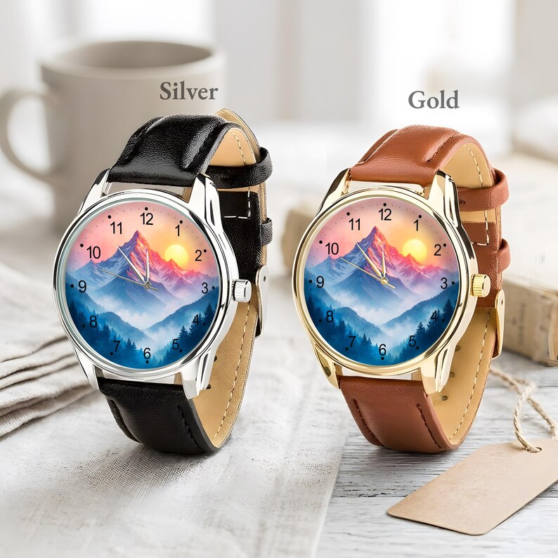 May include: Two wristwatches with mountain landscape dials. One watch has a silver case and black leather strap, the other has a gold case and brown leather strap. The watch faces feature a mountain range with a sunset.
