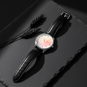 May include: A wristwatch with a black leather strap and a silver-tone case. The watch face features a watercolor-style background in shades of pink, orange, and blue, with black numerals and thin black hands. A delicate botanical illustration is visible.