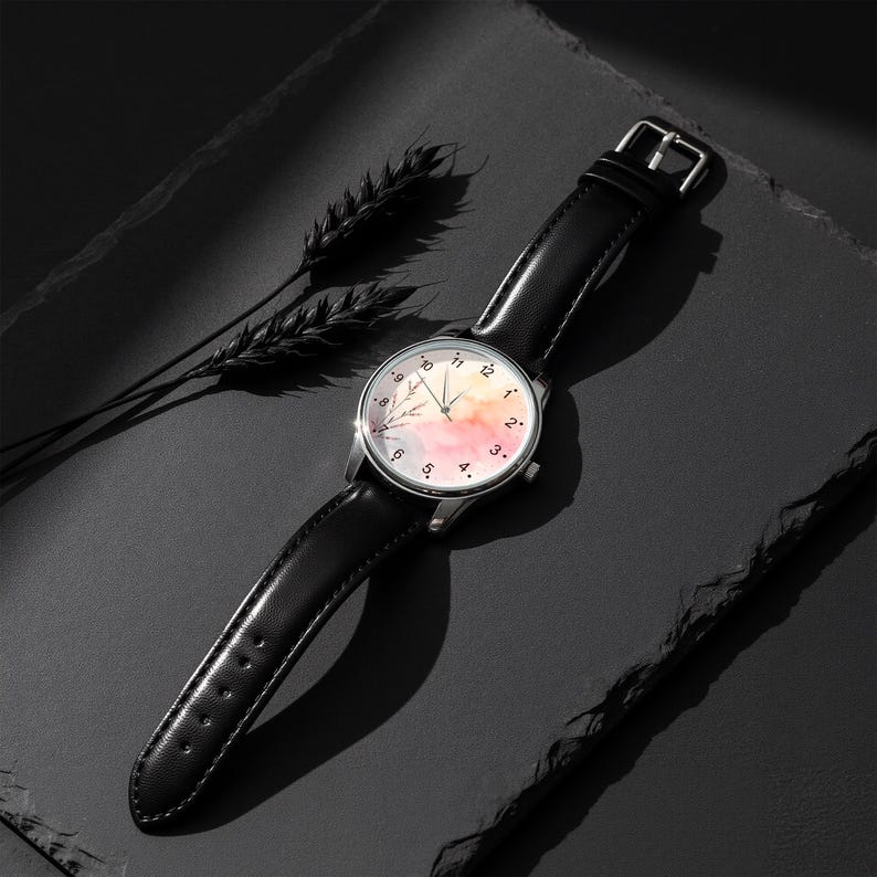 May include: A wristwatch with a black leather strap and a silver-tone case. The watch face features a watercolor-style background in shades of pink, orange, and blue, with black numerals and thin black hands. A delicate botanical illustration is visible.