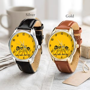 Puede incluir: Dos relojes de pulsera con esferas amarillas que muestran un gr&aacute;fico de cuatro pl&aacute;tanos de dibujos animados con gafas de sol y sombreros. Uno tiene caja plateada con correa de cuero negra y el otro caja dorada con correa de cuero marr&oacute;n.