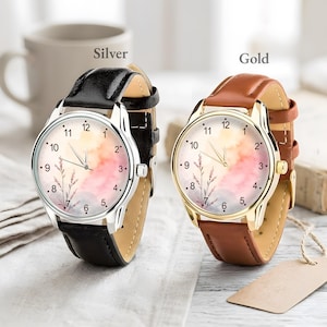 May include: Two analog wristwatches with round faces featuring a watercolor-style background in shades of pink, orange, and blue with a delicate floral illustration. One watch has a silver-tone case and black leather strap, the other a gold-tone case and brown leather strap.