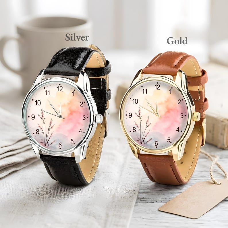 May include: Two analog wristwatches with round faces featuring a watercolor-style background in shades of pink, orange, and blue with a delicate floral illustration. One watch has a silver-tone case and black leather strap, the other a gold-tone case and brown leather strap.