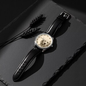 Pu&ograve; includere: Un orologio da polso con cinturino in pelle nera e cassa color argento. Il quadrante presenta un'illustrazione color seppia del volto di un cane, con impronte di zampe sul bordo. L'orologio &egrave; ambientato su uno sfondo scuro.