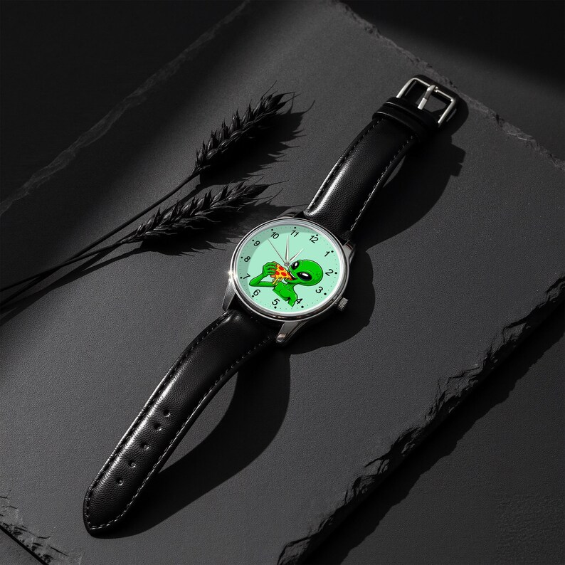 May include: A wristwatch with a black leather strap and a silver-tone metal case. The watch face is light green and features a graphic illustration of a green alien eating a slice of pepperoni pizza, with black numbers marking the hours.