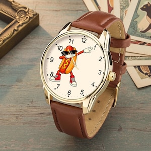May include: A wristwatch with a gold-tone metal case and a brown leather strap. The watch face features a cream background with black numerals from 1 to 12 and a central graphic of a hot dog character wearing sunglasses and red sneakers while dabbing.