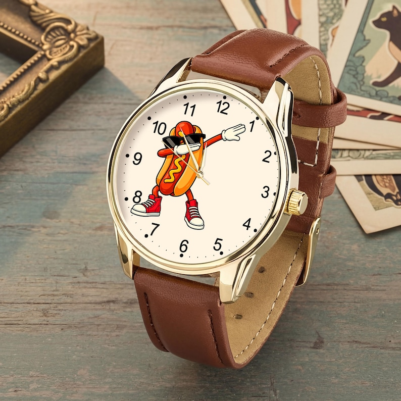 May include: A wristwatch with a gold-tone metal case and a brown leather strap. The watch face features a cream background with black numerals from 1 to 12 and a central graphic of a hot dog character wearing sunglasses and red sneakers while dabbing.