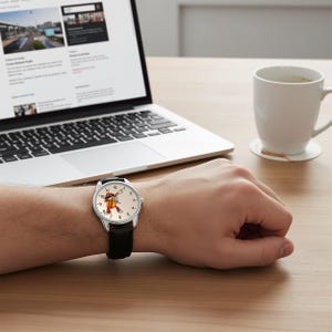 May include: A wristwatch with a silver-tone case and a black leather strap. The watch face is off-white with black numerals and features a graphic illustration of a hot dog character performing a dabbing pose in the center of the dial.