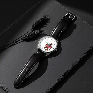 May include: A wristwatch with a black leather strap and a silver-tone circular case. The white watch face features black numerical hour markers and a central graphic of a brown creature wearing a colorful shirt and a pink inflatable pool ring.