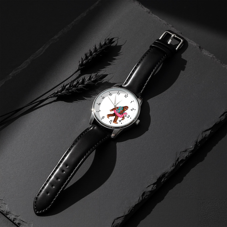 May include: A wristwatch with a black leather strap and a silver-tone circular case. The white watch face features black numerical hour markers and a central graphic of a brown creature wearing a colorful shirt and a pink inflatable pool ring.