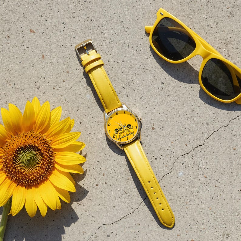 Puede incluir: Un reloj de pulsera amarillo con una correa de cuero amarilla a juego y una esfera redonda con un gr&aacute;fico de cuatro pl&aacute;tanos de dibujos animados con gafas de sol. El reloj est&aacute; colocado junto a un girasol amarillo y gafas de sol.