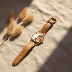 May include: A gold-toned wristwatch with a brown leather band. The watch face features a whimsical design of stacked cats in various colors. The watch is set against a neutral-toned background with dried decorative plants.