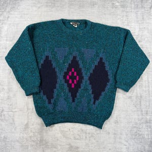 May include: A long-sleeve crew neck sweater in a speckled teal knit. The front features a large, centered dark navy diamond pattern with a smaller, bright magenta geometric design inside the middle diamond, framed by ribbed cuffs and hem.